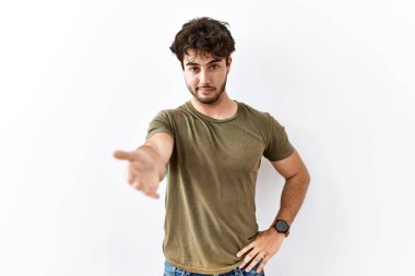 Hispanic man standing over isolated white background smiling cheerful offering palm hand giving assistance and acceptance. 