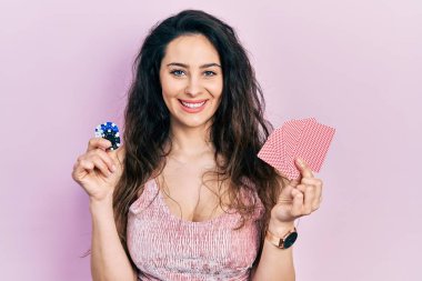 Kumarhane fişleri ve kartlarla poker oynayan genç İspanyol kadın yüzünde mutlu ve havalı bir gülümsemeyle gülümsüyor. diş gösterme. 