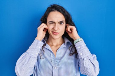 Genç esmer kadın mavi arka planda duruyor. Yüksek sesli müzik sesi için kulaklarını parmaklarıyla kapatıyor. sağır kavramı. 