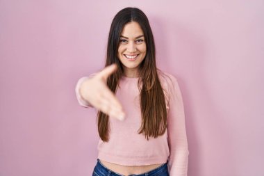 Pembe arka planda duran esmer genç bir kadın selamlama ve hoş geldin demek için dostça bir şekilde tokalaşıyor. başarılı iş. 