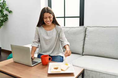 Genç Latin kadın kahvaltısını yapıyor. Evdeki kanepede oturuyor.