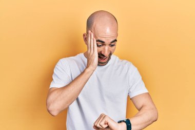 Genç kel adam, günlük beyaz t-shirt giyiyor. Saate bakıyor. Endişeli, geç kalmaktan korkuyor. 