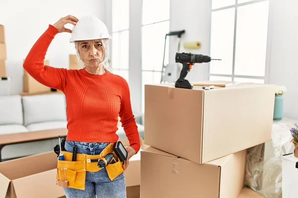 Middle age grey-haired woman wearing hardhat standing at new home confuse and wonder about question. uncertain with doubt, thinking with hand on head. pensive concept.