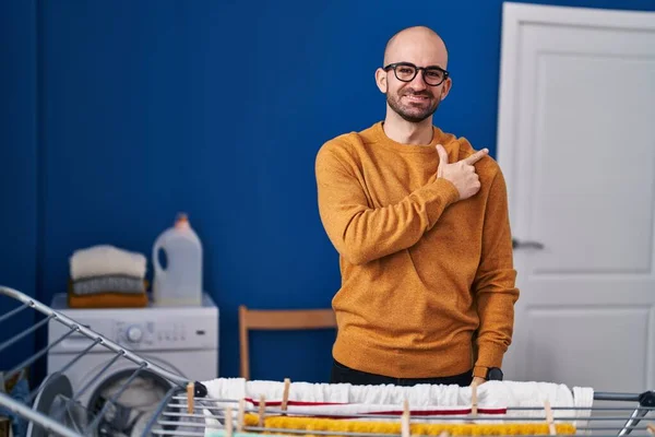 Young bald man with beard hanging clothes at clothesline smiling cheerful pointing with hand and finger up to the side