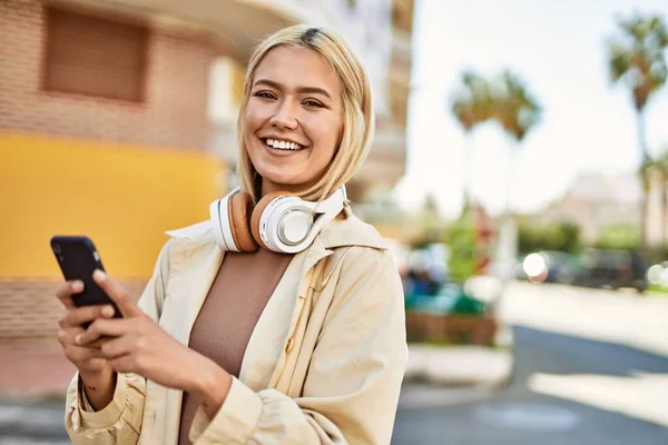 Young blonde girl smiling happy using smartphone and headphones at the city.