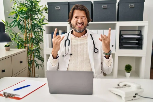 Handsome middle age doctor man working at the clinic shouting with crazy expression doing rock symbol with hands up. music star. heavy music concept.