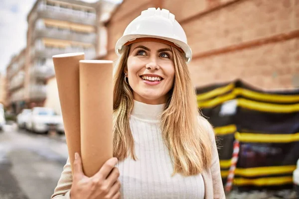 Young blonde woman wearing hardhat holding blueprints at street