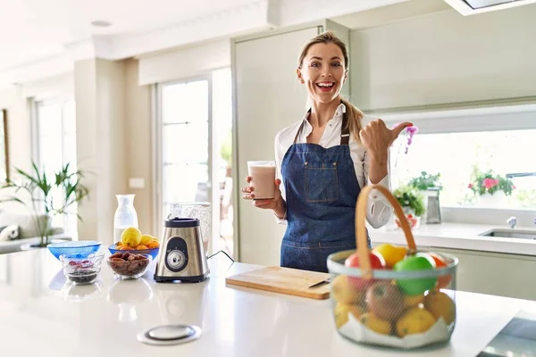 Beautiful Blonde Woman Wearing Apron Drinking Smoothie Kitchen Pointing Thumb — Stok fotoğraf