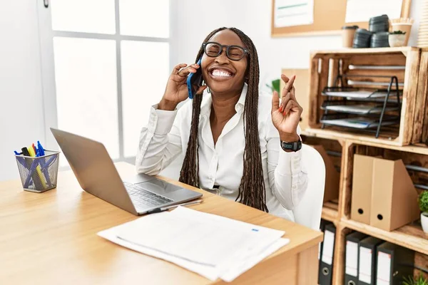 Black woman with braids working at the office speaking on the phone gesturing finger crossed smiling with hope and eyes closed. luck and superstitious concept.