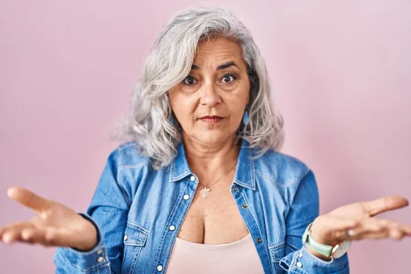 Pembe arka planda duran gri saçlı, kafası karışık, kolları açık orta yaşlı bir kadın.. 