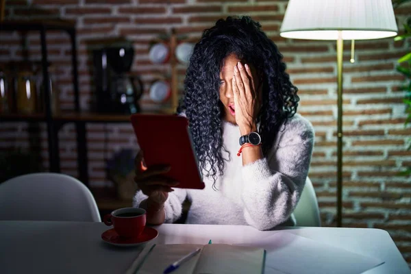 Middle age hispanic woman using touchpad sitting on the table at night yawning tired covering half face, eye and mouth with hand. face hurts in pain.