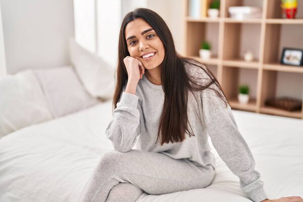 Молодая латиноамериканка улыбается уверенно сидя на кровати в спальне