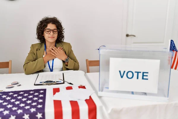 Young hispanic woman at political election sitting by ballot smiling with hands on chest with closed eyes and grateful gesture on face. health concept.