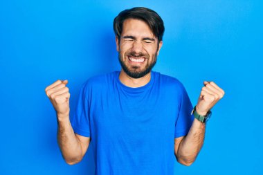 Gündelik kıyafetler giyen genç İspanyol adam çok mutlu ve heyecanlı. Kazanan jestler yapıyor. Kolları havada, gülümsüyor ve başarı için bağırıyor. kutlama konsepti. 