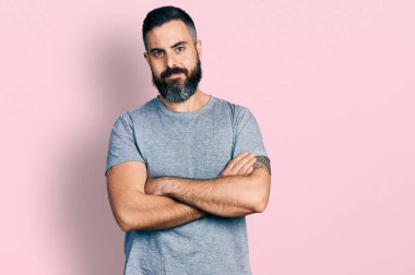 Sakallı, sıradan gri tişörtlü, mutlu yüzlü, kollarını kavuşturup kameraya bakan İspanyol bir adam. pozitif kişi. 