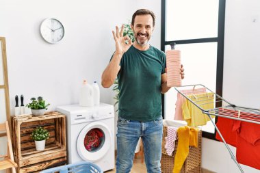 Sakallı orta yaşlı bir adam çamaşır deterjanı şişesini tutuyor. Tamam işareti yapıyor, gülümsüyor ve mükemmel bir sembol oluşturuyor. 