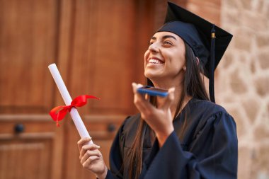 Young hispanic woman wearing graduated uniform holding diploma talking on the smartphone at university