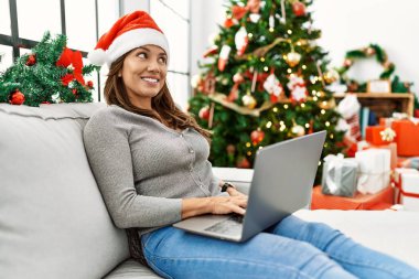 Evde dizüstü bilgisayar kullanan genç Latin kadın Noel ağacının yanında oturuyor.