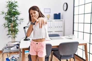 Young hispanic girl standing at pediatrician clinic laughing at you, pointing finger to the camera with hand over mouth, shame expression 