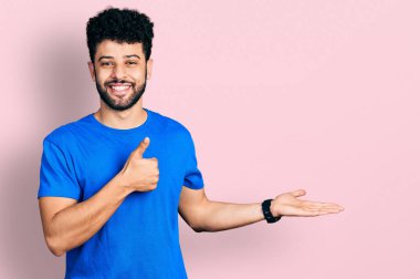 Sakallı, günlük mavi tişörtlü genç bir Arap. Avuç içini gösteriyor ve baş parmaklarıyla hareket ediyor. Mutlu ve neşeli gülümsüyor. 