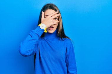 Balıkçı yaka kazak giyen genç esmer kız şok içinde yüzünü ve gözlerini kapayarak utangaç bir ifadeyle parmaklarının arasından bakıyor.. 