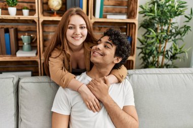 Mutlu gülümseyen ve sarılan genç çift evdeki kanepede oturuyor..