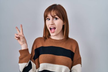 İzole edilmiş arka planda çizgili süveter giyen genç ve güzel bir kadın kameraya gülümseyen bir gülümsemeyle parmaklarıyla zafer işareti yapıyor. İki numara.. 