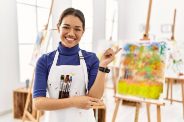 Sanat stüdyosundaki genç esmer kadın yüzünde büyük bir gülümsemeyle, kameraya el ile işaret ederek bakıyor.. 