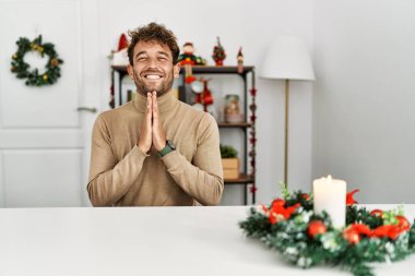 Genç, sakallı yakışıklı adam Noel süslemesi masasında oturmuş elleriyle dua ederek af diliyor kendinden emin bir şekilde gülümsüyor.. 
