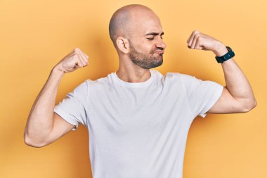 Sıradan beyaz tişört giyen genç kel adam gururla gülümseyen kol kaslarını gösteriyor. fitness konsepti. 