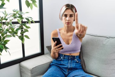 Young beautiful woman using smartphone typing message sitting on the sofa pointing with finger up and angry expression, showing no gesture 