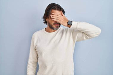 Mavi arka planda sıradan süveter giyen yakışıklı orta yaşlı bir adam. Gülümsüyor ve elleri yüzünde, şaşırmak için gözlerini kapatıyor. kör kavram. 