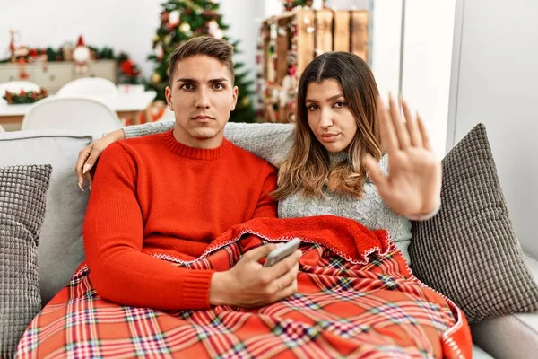 Young hispanic couple sitting on the sofa on christmas with open hand doing stop sign with serious and confident expression, defense gesture