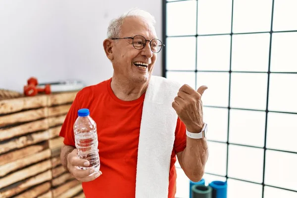 Senior man wearing sportswear and towel at the gym smiling with happy face looking and pointing to the side with thumb up.