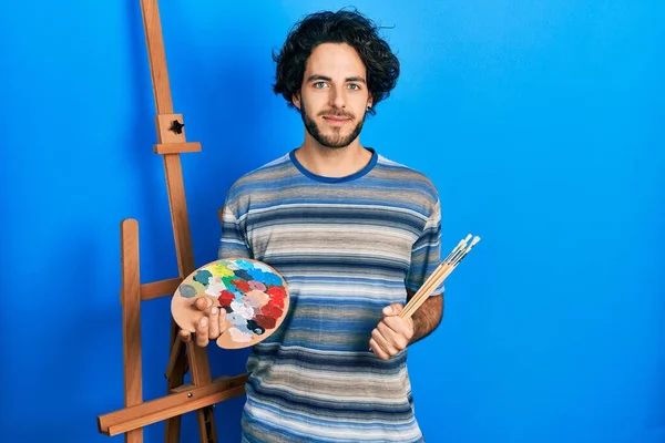 Handsome hispanic man standing drawing with palette by painter easel stand relaxed with serious expression on face. simple and natural looking at the camera.
