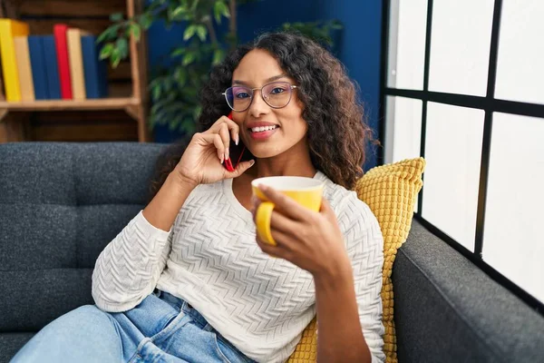 Genç Latin kadın kahve içiyor ve evde akıllı telefondan konuşuyor.