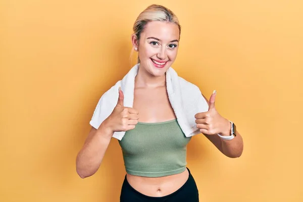 Young blonde girl wearing sportswear and towel success sign doing positive gesture with hand, thumbs up smiling and happy. cheerful expression and winner gesture.