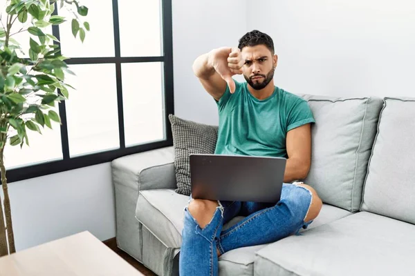 Young handsome man with beard using computer laptop sitting on the sofa at home looking unhappy and angry showing rejection and negative with thumbs down gesture. bad expression.