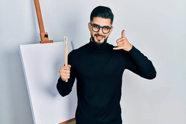 Handsome man with beard holding brushes close to easel stand smiling doing phone gesture with hand and fingers like talking on the telephone. communicating concepts.