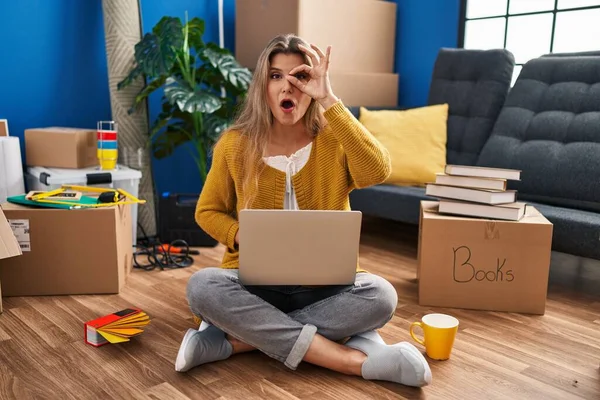 Young woman sitting on the floor at new home using laptop doing ok gesture shocked with surprised face, eye looking through fingers. unbelieving expression.