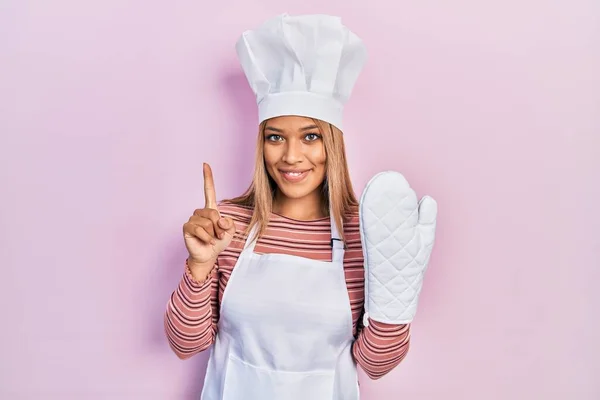 Beautiful hispanic woman uniform wearing apron and hat wearing protective glove smiling with an idea or question pointing finger with happy face, number one