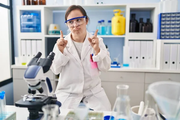 Hispanic girl with down syndrome working at scientist laboratory pointing up looking sad and upset, indicating direction with fingers, unhappy and depressed.