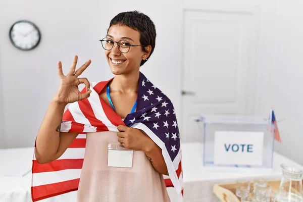 Young hispanic woman with short hair at political campaign election holding usa flag doing ok sign with fingers, smiling friendly gesturing excellent symbol