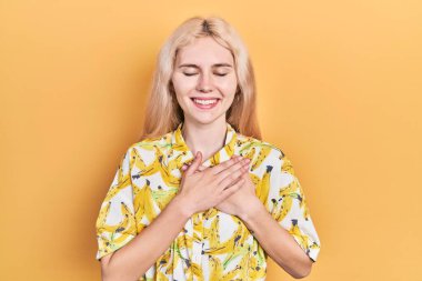 Beautiful caucasian woman with blond hair wearing colorful shirt smiling with hands on chest with closed eyes and grateful gesture on face. health concept. 