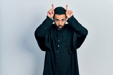 Sakallı, Katolik papaz cüppeli yakışıklı bir adam boynuzları gibi parmağıyla komik hareketler yapıyor. 