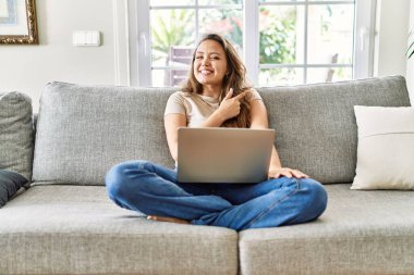 Güzel, genç, esmer bir kadın evinde bilgisayar laptopuyla oturuyor. Yüzünde mutlu ve doğal bir ifadeyle, elleri ve parmaklarıyla gülümsüyor. 