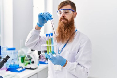 Laboratuvarda test tüpleri tutan bilim adamı üniforması giyen genç kızıl saçlı adam.