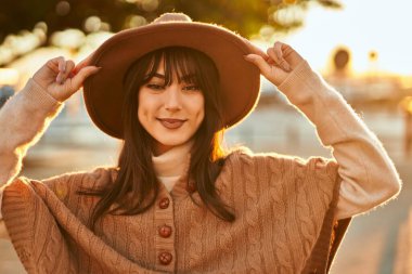 Kışlık şapkalı esmer kadın gün batımında şehirde gülümsüyor.