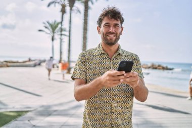 Genç İspanyol adam deniz kenarında akıllı telefon kullanarak gülümsüyor.
