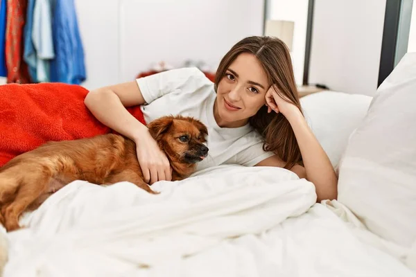 Young hispanic woman hugging dog lying on bed at bedroom